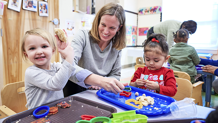 children playing with Play-Dough and cookie cutters with a smiling teacher